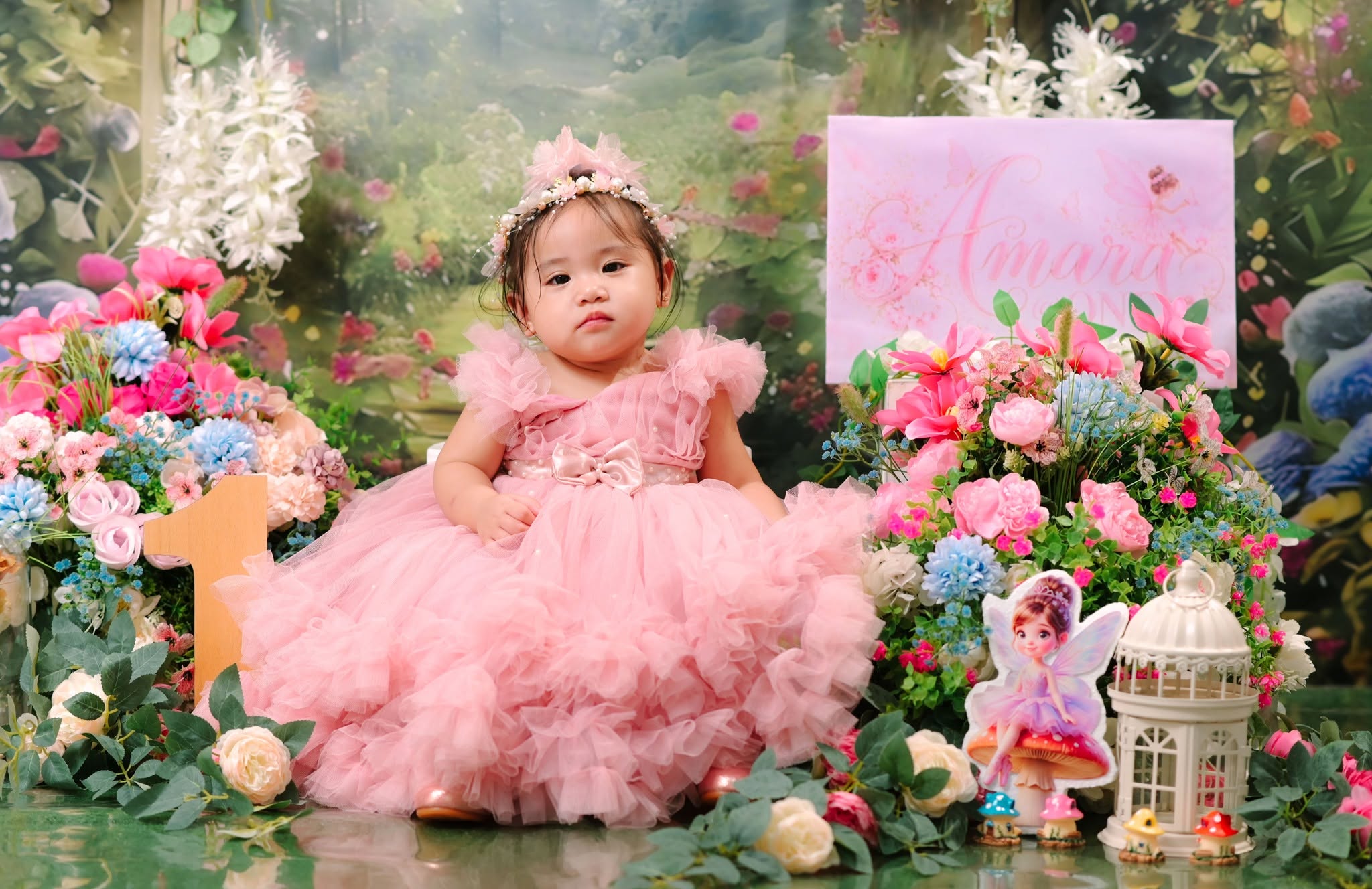 Amara seated in her pink birthday dress among flowers, beside a number one and fairy decorations